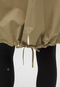 Khaki bunda s staženým lemem, zdobená mašlí a bočními rozparky. Pod ní jsou viditelné černé legíny.