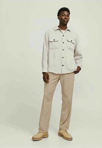 Young man stands wearing beige button-up shirt with chest pockets, light tan pants, and beige lace-up shoes against a neutral background.