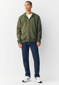 Junger Mann steht, trägt einen olivgrünen Reißverschluss-Hoodie, ein weißes Shirt, dunkelblaue Jeans und weiße Turnschuhe mit schwarzen Akzenten.