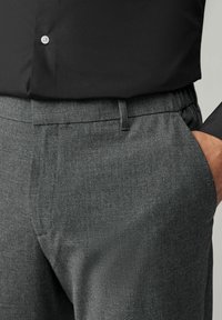 Graue gewebte Hose mit flachem Frontschnitt, seitlichen Taschen und elastischem Bund. Kombiniert mit einem schwarzen Hemd mit Knöpfen.