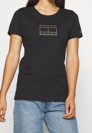 Femme portant un t-shirt noir Tommy Jeans avec un logo doré et un jean en denim bleu clair sur un fond uni.