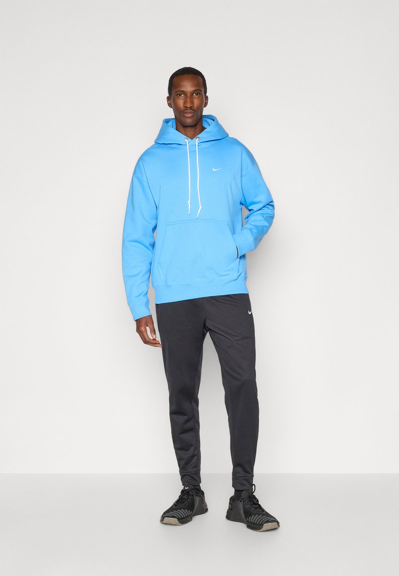 Helle blaue Kapuzenjacke mit einer Vordertasche und weißem Kordelzug, kombiniert mit dunkelgrauen, schmal geschnittenen Hosen und schwarzen Sportschuhen.