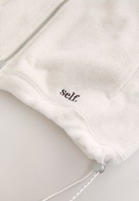 Veste en polaire blanche avec fermeture éclair, cordon de serrage à l’ourlet et "self." brodé en noir près du bord inférieur.