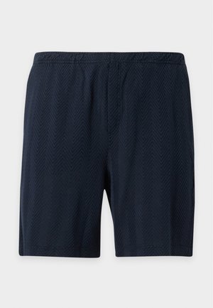 Hombre que lleva una camiseta blanca con diseño cuadrado ondulado y texto, pantalones cortos azul marino texturizados y mocasines negros, de pie frente a un fondo blanco.