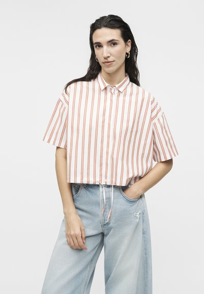 Γυναίκα με μακριά σκουρόχρωμα μαλλιά φοράει χρυσούς κρίκους σκουλαρίκια, κοντό λευκό πουκάμισο με κάθετες πορτοκαλί ρίγες και ανοιχτό μπλε τζιν παντελόνι με φαρδιές παντελόνια ψηλόμεσο.