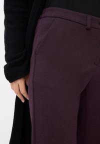 Dunkellila strukturierte Hose mit geometrischem Muster, seitlichen Taschen und taillierter Passform, ergänzt durch ein schwarzes Kleidungsstück.