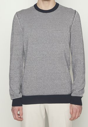 Mann trägt einen langärmligen Pullover mit marineblau-weißem Hahnentrittmuster, mit marineblauen Bündchen, Kragen und Saum, kombiniert mit hellen Hosen.