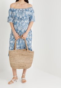 Kudottu olkakassi, jossa on paksut kahvat, beige väri ja tekstuuri; yhdistetty siniseen kukalliseen olkaimettomaan mekkoon ja valkoisiin sandaaleihin.