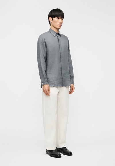 Γκρι πουκάμισο με μακριά μανίκια και κουμπιά, κατασκευασμένο από ελαφρύ ύφασμα, με γιακά και ίσια γραμμή, συνδυασμένο με κρεμ παντελόνια και μαύρα παπούτσια.