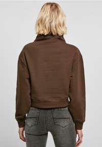 Sudadera marrón recortada con cuello alto, textura suave y diseño de hombros caídos, combinada con vaqueros ajustados gris oscuro.