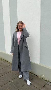 Cappotto grigio doppiopetto con rever a punta sopra una camicia rosa con bottoni, abbinato a pantaloni grigi a gamba larga e sneakers bianche.