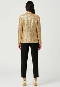 Blazer in paillettes dorées con un design aderente, stile monopetto e rever a punta, abbinato a pantaloni neri su misura e tacchi dorati.
