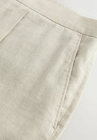 Pantalons en mélange de coton beige avec une finition texturée, dotés d'une poche latérale et de détails de couture subtils le long des bords.