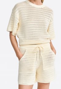 Vaatteet beigen värin neulepuuvilla loungewear-setti, joka sisältää lyhythihaisen paidan pyöreällä kaula-aukolla ja siihen sopivat shortsit, joissa on elastinen vyötärö ja taskut.