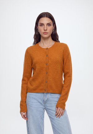 Femme aux longs cheveux foncés porte un cardigan orange boutonné et un jean bleu clair, debout devant un fond blanc uni.