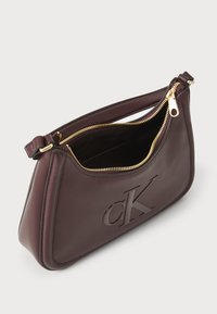 Bordeaux leren handtas met een gegraveerd "CK" logo, ritsluiting en goudkleurige hardware. Heeft een ronde vorm en een gladde textuur.