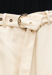 Detailný záber na béžový látkový opasok so zlatou kovovou prackou a očkami, zapnutý okolo pása, nad ktorým je vidieť čierny rebrovaný top.