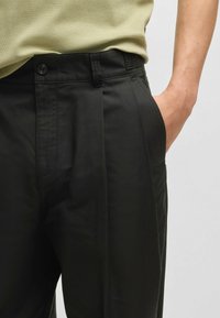 Pantalons noirs plissés avec une texture lisse, dotés de poches latérales et d'une fermeture à bouton. Associés à un haut kaki clair.