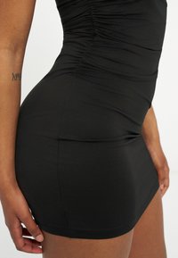 Gros plan d'une personne portant une robe noire cintrée à fronces, montrant le côté du torse et le haut de la cuisse avec un tatouage visible sur le bras.