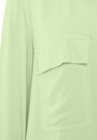 Light green, woven fabric with a smooth texture; features a chest pocket and stitched accents. Close-up of the garment.