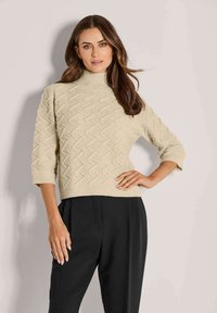 Frau mit langen braunen Haaren, die einen beigefarbenen strukturierten Rollkragenpullover mit Dreiviertelärmeln und hochtaillierten schwarzen Hosen trägt und steht.