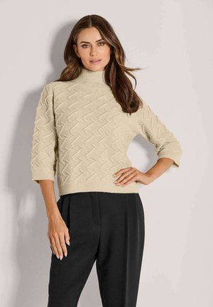 Vrouw met lang bruin haar, gekleed in een beige textuur turtleneck trui met driekwart mouwen en hooggemaakt zwarte broek, staande.