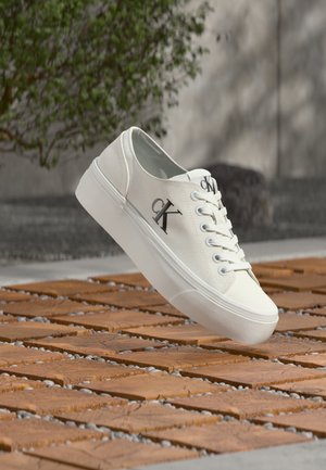 Bijela Calvin Klein tenisica s platformom potplatom lebdi iznad drvenog pločnika na otvorenom, u blizini zelenog grmlja.