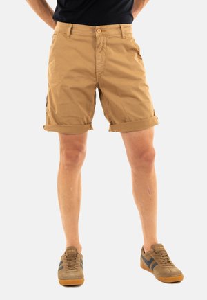 Shorts en coton beige avec des poignets roulés, des poches avant et une fermeture à bouton. Portés avec des sneakers marron ornés de rayures foncées.