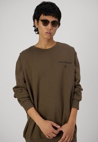 Felpa marrone con maniche lunghe, polsini a coste e logo "Lunar Hypnosis" sul petto. Indossata con occhiali da sole ovali.