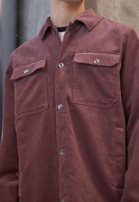 Bruine corduroy shirt met twee borstzakken, knoopsluitingen en een puntige kraag. Beschikt over een subtiele ribbeltjesstructuur en metalen knopen.