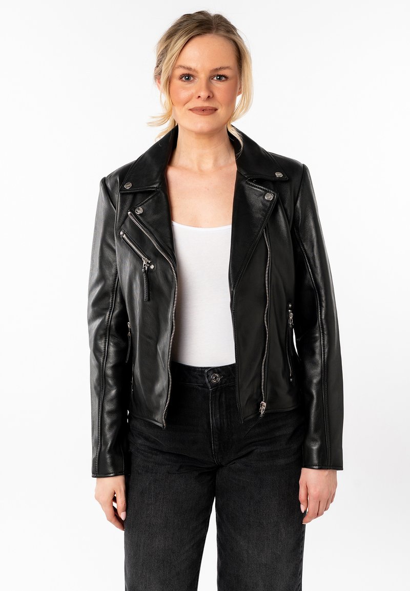 Giacca di pelle nera da motociclista con colletto a REVER, zip asimmetrica e tasche con zip. Indossata sopra a una maglietta bianca e jeans neri.