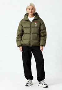 Veste matelassée vert olive avec fermeture éclair devant, capuche et poignets côtelés ; présente un motif blanc de main et de cœur sur la poitrine. Pantalon cargo noir et baskets.