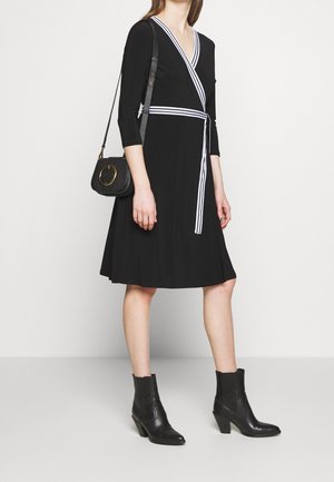 Vestito avvolgente nero con strisce bianche, maniche a tre quarti, tessuto morbido, gonna fino al ginocchio. Abbinato a stivaletti neri con tacco e una piccola borsa.