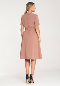 Różowa sukienka z krótkimi rękawami, dopasowanym wcięciem w talii i spódnicą w kształcie litery A, z zapięciem z tyłu, noszona z czarnymi szpilkami.