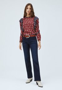 Blusa rossa con motivo paisley e rifiniture volants in blu navy, indossata con jeans scuri e una cintura marrone. L'outfit è completato da stivaletti alla caviglia color crema.