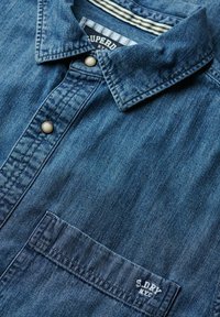 Denim shirt in donkerblauw, met een button-down kraag, een voorzak en geborduurde branding. Zachte textuur met een lichte vervaging.