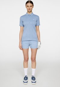 Vaaleansininen lyhythihainen polo-paita, jossa on hienovarainen tekstuuri, yhdistettynä samanvärisiin shortseihin. Valkoiset hanskat ja urheilulliset siniset lenkkarit täydentävät asukokonaisuuden.