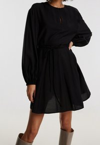 Robe noire à col montant, manches longues bouffantes et ceinture à la taille. Comprend une découpe centrale en forme de goutte et un ourlet évasé.
