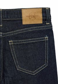 Jeans en denim bleu foncé avec une pièce en cuir portant le logo "VERO MODA" cousu à l'arrière de la taille. Des surpiqûres jaunes contrastent avec le tissu.