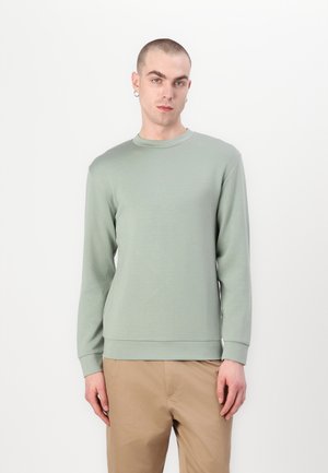 Light green long-sleeve sweatshirt, soft fabric, crew neck, ribbed cuffs and hem, paired with light brown trousers. Minimalist design.