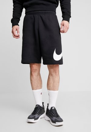 Mężczyzna w czarnych spodenkach Nike z dużym białym logo, czarnych tenisówkach, białych skarpetkach i czarnej bluzie, stojący na szarej podłodze.