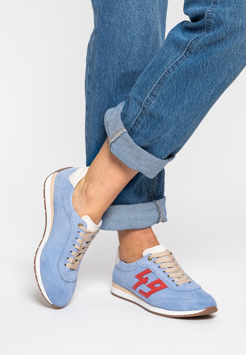 Personne portant un jean bleu retroussé et des baskets bleu clair avec des lacets beige et un détail rouge "49" sur fond blanc.