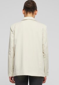 Blazer crema con vestibilità su misura, revers a punta, maniche lunghe e una texture liscia. La vista posteriore mostra un design pulito e minimalista.