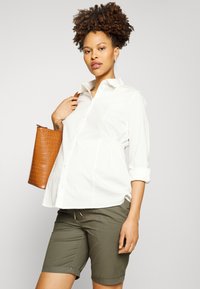Camisa branca de botão com mangas dobradas, apresenta detalhes franzidos nas laterais. Combinada com calções verdes-oliva e uma mala tote castanha com padrão de croco.