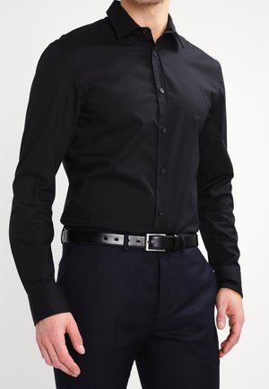 Homme portant une chemise noire ajustée boutonnée rentrée dans un pantalon sombre avec une ceinture en cuir noire, debout devant un fond uni.