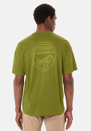 Man draagt een olijfgroen T-shirt met een geel grafisch ontwerp van een berg, zon en fiets op de achterkant, gecombineerd met beige broek.