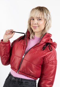 Giacca puffer rossa corta con una texture lucida, cordoncini neri sul cappuccio e una zip frontale. Indossata sopra un maglione rosa.