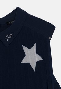 Chemise bleue marine avec un col pointu, présentant un grand motif d'étoile blanche et des coutures argentées sur le col. Comprend des accents en matériel argenté.