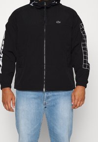 Svart vindjacka med dragkedja framtill, justerbar huva, kontrasterande vit logotyp och rutmönster längs ärmarna, samt elastiska ärmslut.