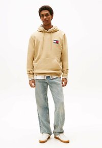Beige hættetrøje med frontlomme og snor i hætten, prydet med et multikolor logo patch. Båret med lyseblå lige ben jeans og hvide sneakers.
